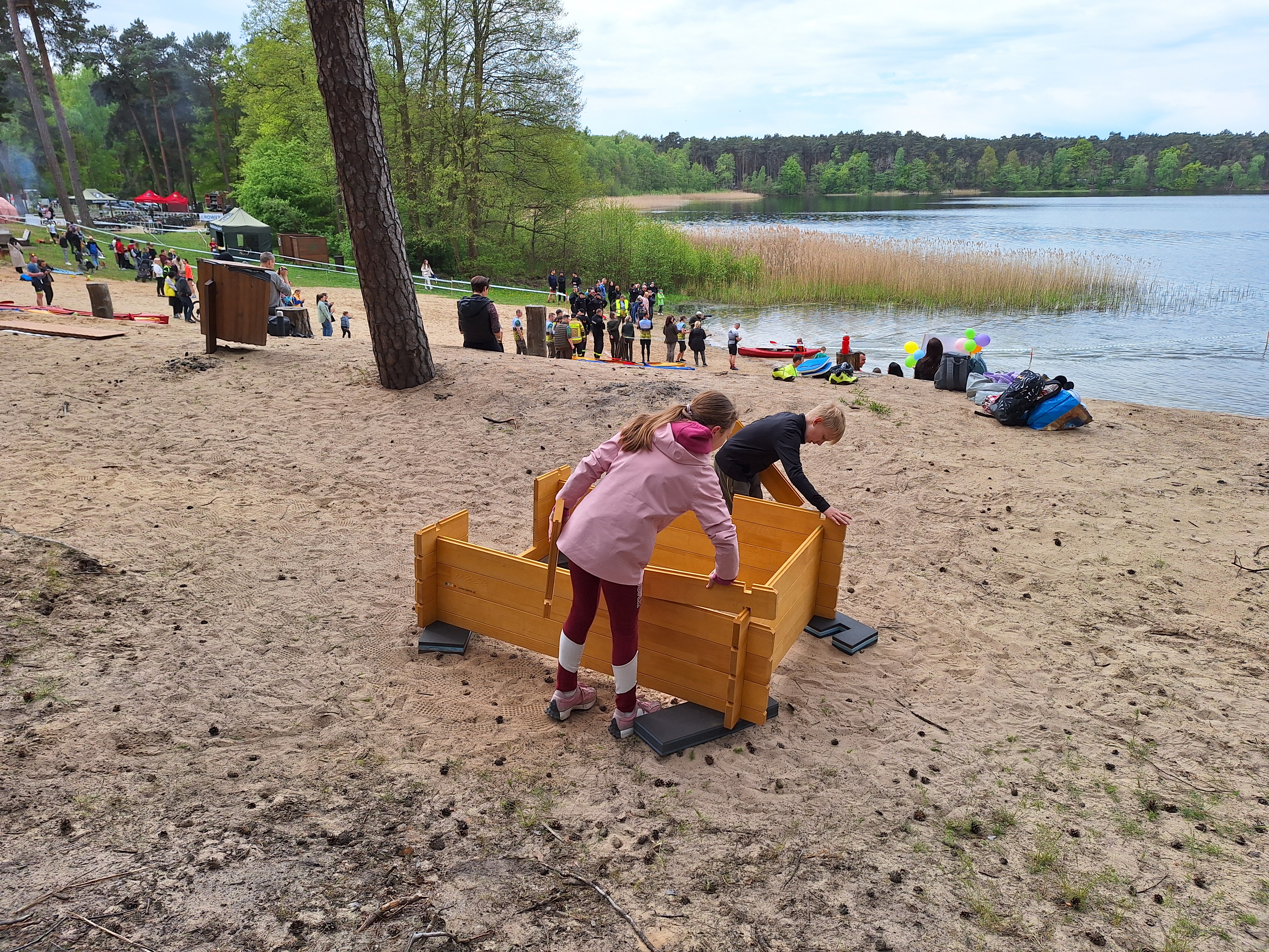 Zdjęcie przedstawia dzieci składające drewniany model domu, w tle jezioro i uczestnicy zawodów na brzegi. Fot. Archiwum N. Grodzisk