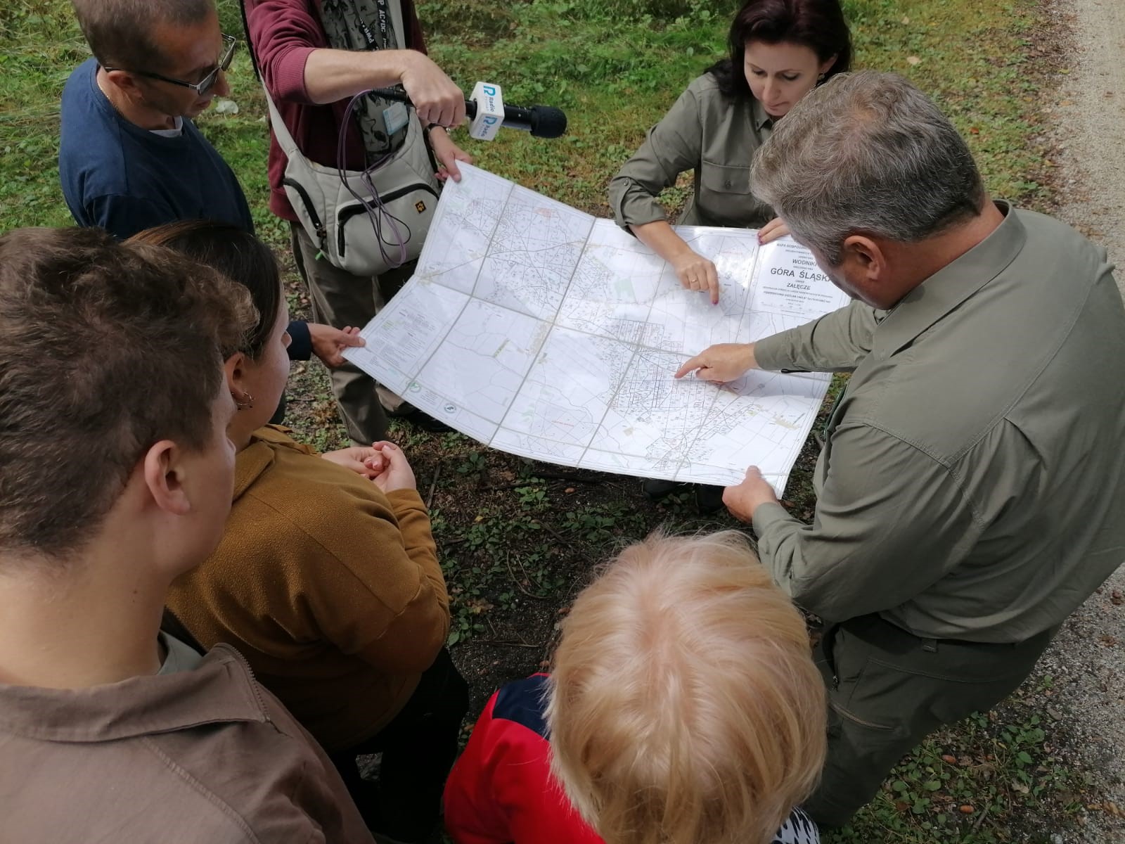 Zdjęcie przedstawia ludzi zgromadzonych wokół mapy oraz redaktorem z mikrofonem nagrywającym ich rozmowę w lesie. Fot. Archiwum RDLP w Poznaniu 