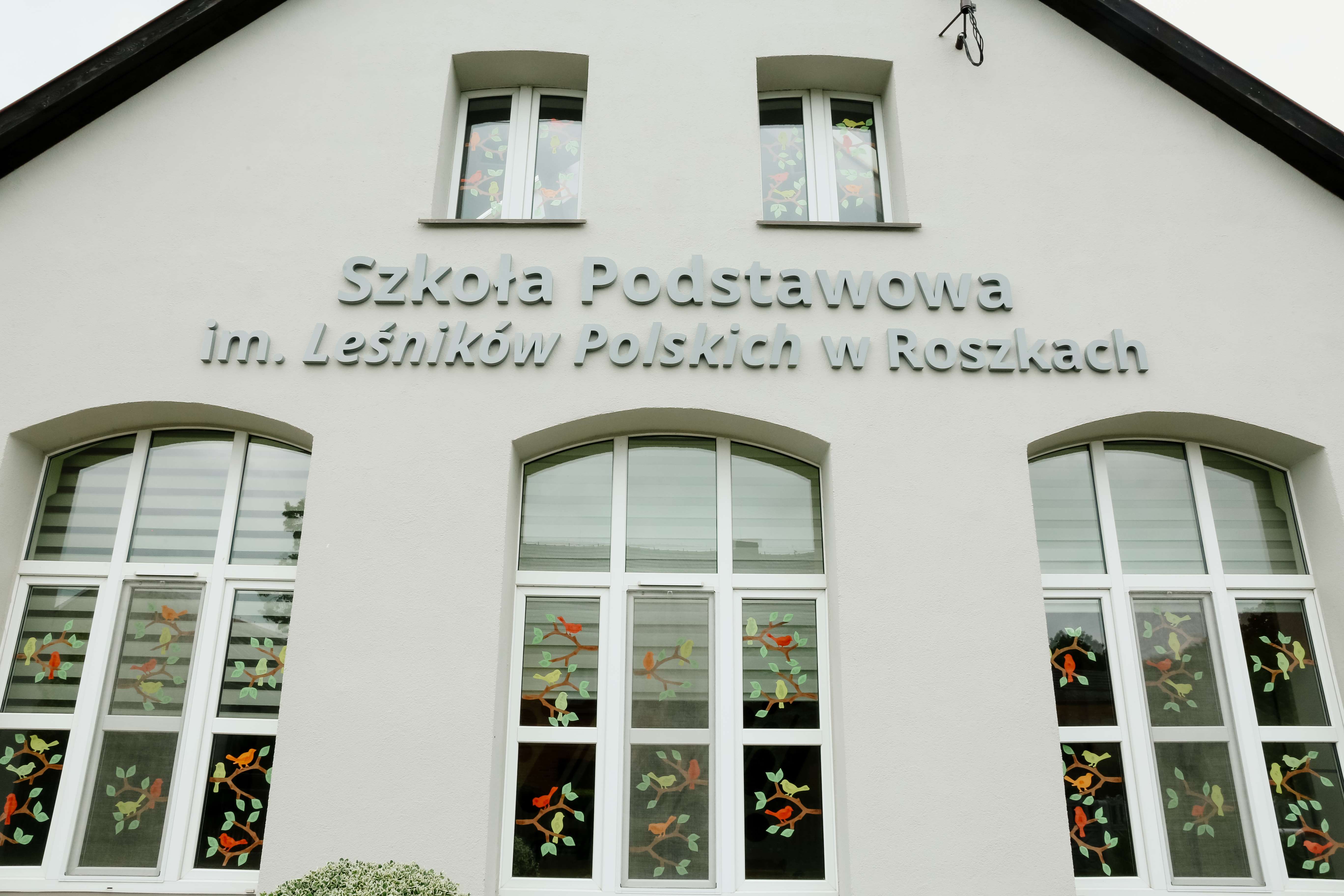Zdjęcie przedstawia zewnętrzną ścianę budynku szkoły z pełną nazwą i oknami, w których widać wycięte z papieru gałęzie drzew i ptaki. Źródło: Archiwum SP w Roszkach